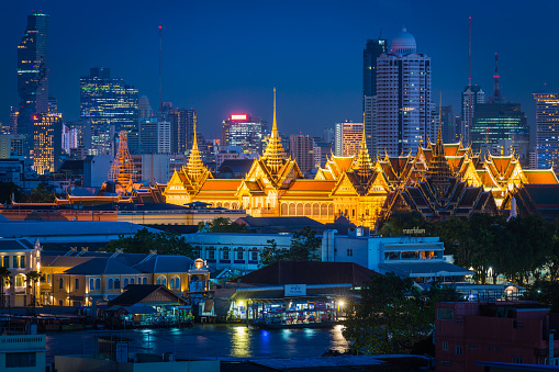 Aerial view over the Chao Phraya River and the spotlit spires of the Grand Palace, temples and futuristic skyscrapers in the heart of Bangkok, Thailand’s vibrant capital city.