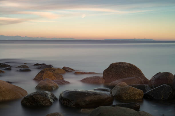 Long exposure of Wet rocks on shore with calm water with mountains and clouds in the distance