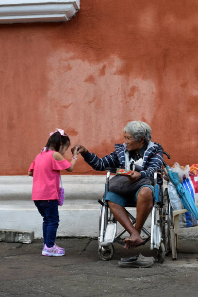 Little girl kisses hand of  Ethnic old woman sitting on wheelchair holding Christmas gift box who begs for alms at old church yard San Pablo City, Laguna, Philippines - December 25, 2018: Little girl kisses hand of  Ethnic old woman sitting on wheelchair holding Christmas gift box who begs for alms at old church yard beg alms stock pictures, royalty-free photos & images