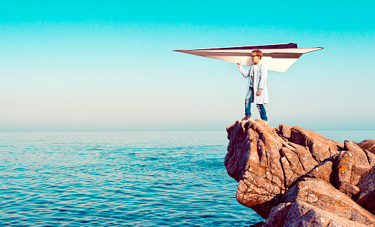 Boy wears a lab coat and stands on a cliff over looking the sea. He holds on to a large paper airplane and he is ready to let it take off. Concept of dreaming big and using imagination to reach your goals.