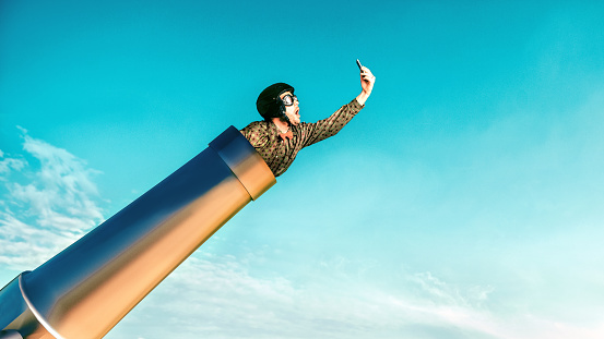 Male dare devil wears helmet and has crawled into a big cannon. He is ready to be shot to the sky and looks at his mobile phone. Concept of getting a fast 4G or 5G connection on your smart phone.