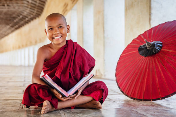 Positivity Novice Monk in Monastery Archway, Myanmar Happy smiling burmese buddhist novice monk sitting on the floor of an archway of buddhist monastery, holding and reading a book, looking up with a bright happy confident smile. Real People. Old Bagan, Mandalay Region, Myanmar. buddhist house of worship stock pictures, royalty-free photos & images