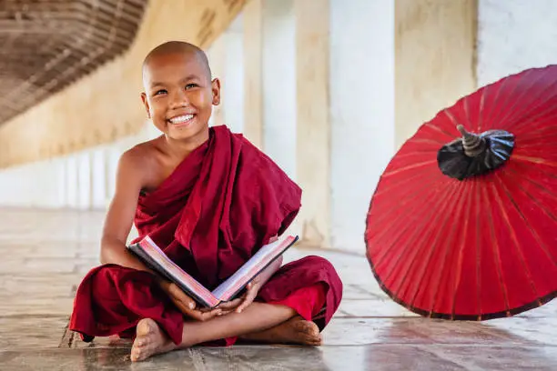 Happy smiling burmese buddhist novice monk sitting on the floor of an archway of buddhist monastery, holding and reading a book, looking up with a bright happy confident smile. Real People. Old Bagan, Mandalay Region, Myanmar.