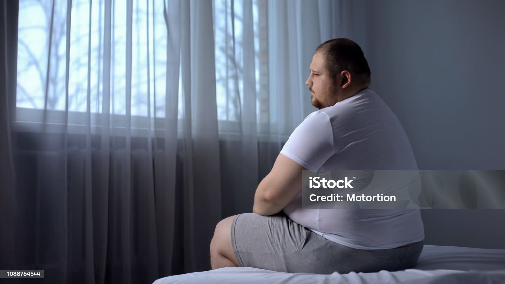 Depressed fat man sitting on bed at home, worried about overweight, insecurities Overweight Stock Photo