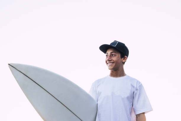 schönes portrait junge teenager mit weißen surfbrett und t-shirt lächelnd und mit blick auf seine seite - weißen himmelshintergrund, für männliche surfer sportler - meer leben lebensstil leute - bali male beautiful ethnicity stock-fotos und bilder