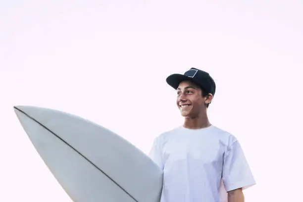 Photo of Nice portrait of young teenager with white surf board and t-shirt smiling and looking at his side - white clear sky background, for male surfer athlete - sea life lifestyle people