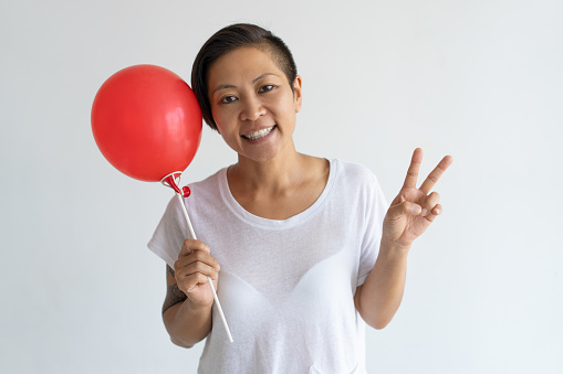 Playful Asian woman holding red balloon and showing victory sign. Smiling lady looking at camera. Party concept. Isolated front view on white background.