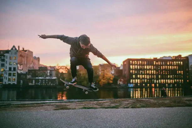 Young man skateboarding in Berlin by the Spree river. He is wearing casual skateboarding clothing, a hoodie and skate shoes, practicing kickflip, ollie and other tricks. Taken on a nice Autumn day, just as the sun sets in Berlin's Friedrichshain - Kreuzberg district near the remaining parts of the Berlin Wall.