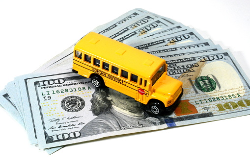 Yellow school bus on top of $100 dollar bills against a white background.