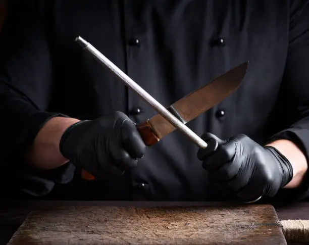 cook in black latex gloves sharpens a knife over a wooden table