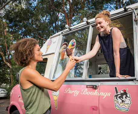 Smiling young Caucasian female ice cream vendor in pink retro style ice cream van sells chocolate-topped ice cream cone to happy mixed race young woman wearing tank top. The suntanned customer has an Afro hairstyle.