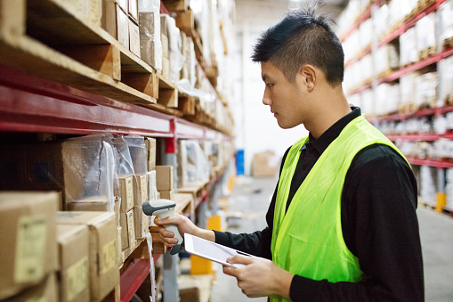 Warehouse worker checking cargo on shelves with scanner. Male worker in uniform holding digital tablet and scanning boxes in shelves.