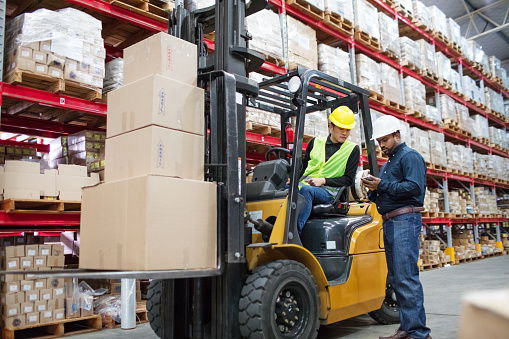 Young man moving load with forklift loader in warehouse and talking with supervisor. Supervisor explaining the cargo details with forklift operator.