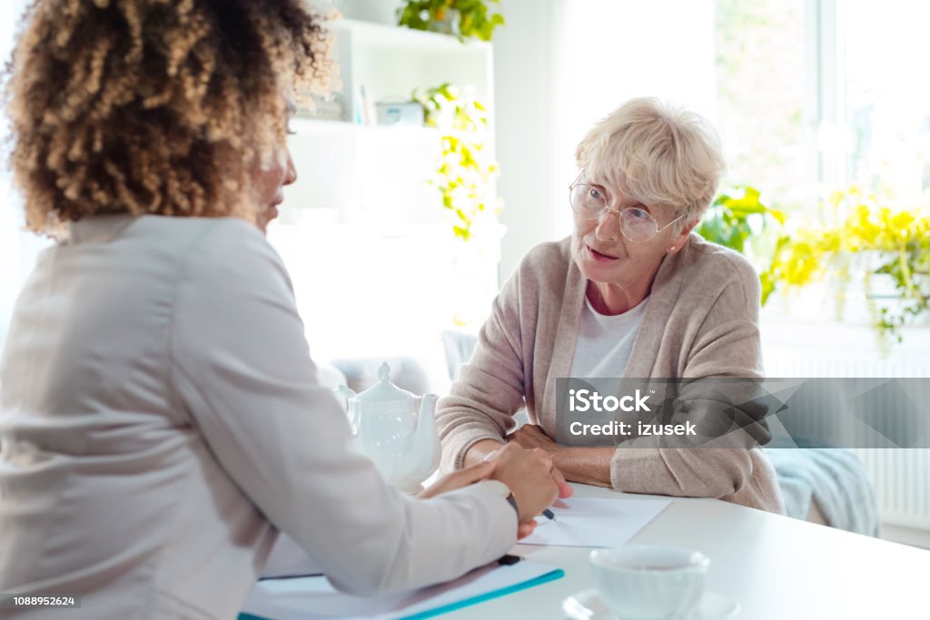 Senior woman talking with insurance advisor Worried senior woman talking with financial advisor, sitting at the table in living room together. Assistance Stock Photo