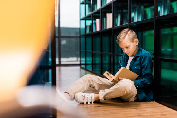 istock cute focused boy reading book and sitting on floor in library 1088955278