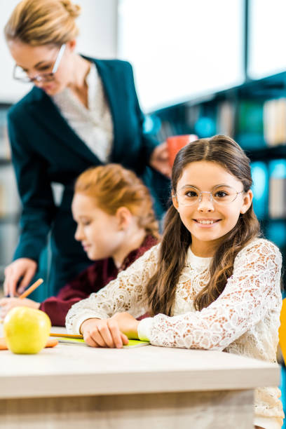 istock cute schoolkid in eyeglasses smiling at camera while studying with classmate and teacher in library 1088955426