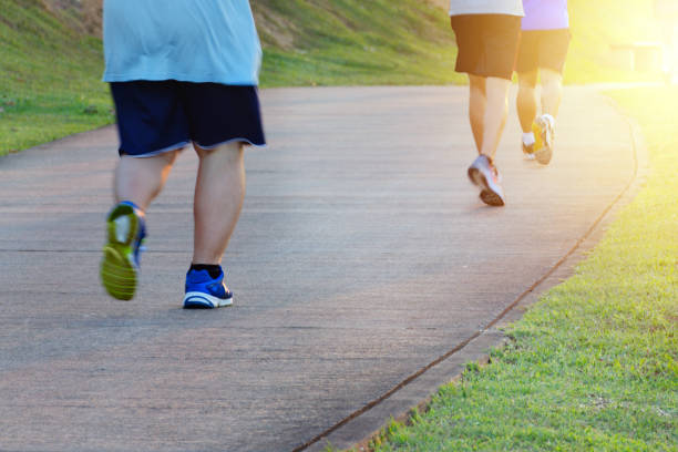 Fat man jogging, catching up with thin men.  Fitness and healthy lifestyle, outdoor sports activity Fat man jogging, catching up with thin men. Low angle view of runners running in park. Blurred feet motion group of runners, Fitness and healthy lifestyle, outdoor sports activity. Copy space. running jogging men human leg stock pictures, royalty-free photos & images