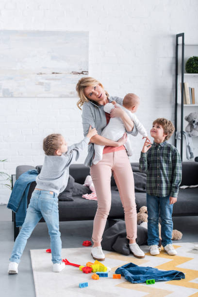 istock exhausted mother talking by smartphone and holding infant child while siblings playing in room 1088961262