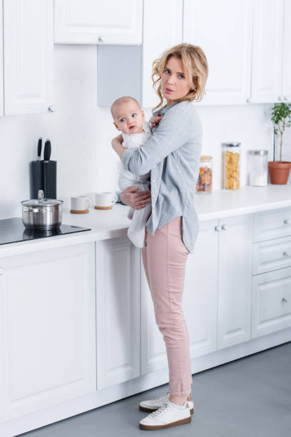 istock tired mother holding infant baby and looking at camera in kitchen 1088961376
