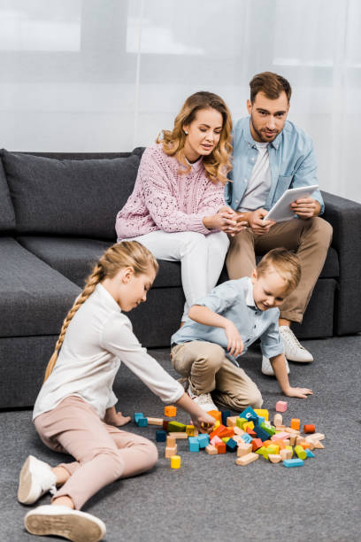 istock parents sitting on sofa with digital tablet and looking at children playing with multicolored wooden blocks on floor in apartment 1088970982