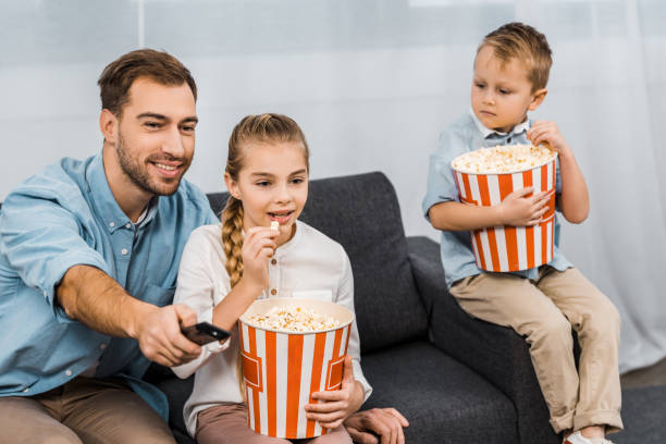 istock smiling father sitting on sofa and changing channels by remote controller with children holding striped buckets and eating popcorn in apartment 1088971088