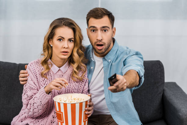istock open-eyed man sitting on sofa, holding remote controller and embracing surprised wife with striped popcorn bucket 1088971106
