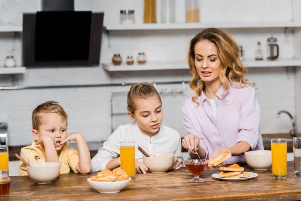 istock cute siblings looking at pretty woman spreading jam on toast at table 1088971232