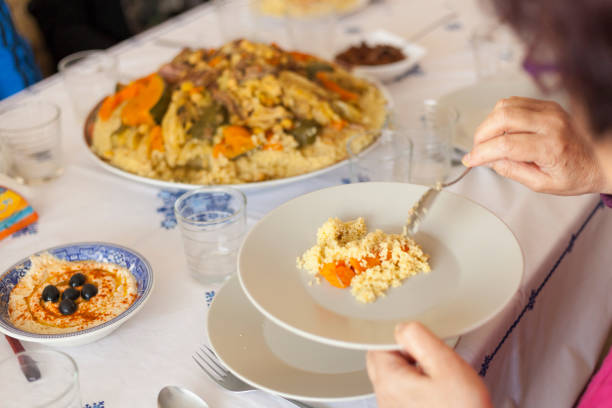 A large plate of typical Moroccan couscous in the centre of a table, napkins, hummus with black olives, glasses and out of focus hand serving some semolina and carrots on a plate.