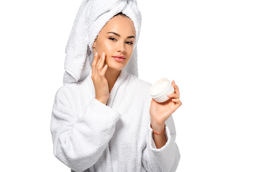beautiful girl in bathrobe looking at camera, holding jar and applying cream on face isolated on white