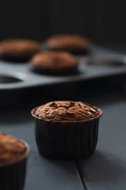 Minimalist style hygge dessert. Chocolate muffins with chocolate drops in brown paper and in baking tray on dark background vertical