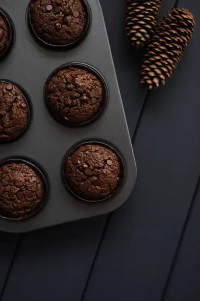 Hygge homemade sweets. Chocolate muffins with chocolate drops in baking tray and dry cones on dark background top view copyspace