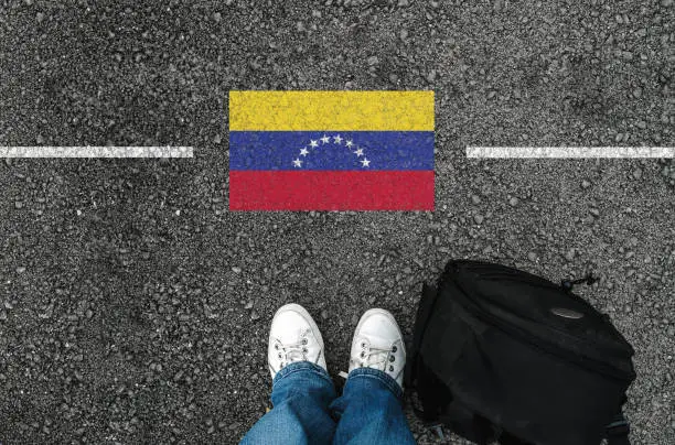 a man with a shoes and backpack is standing on asphalt next to flag of Venezuela and border