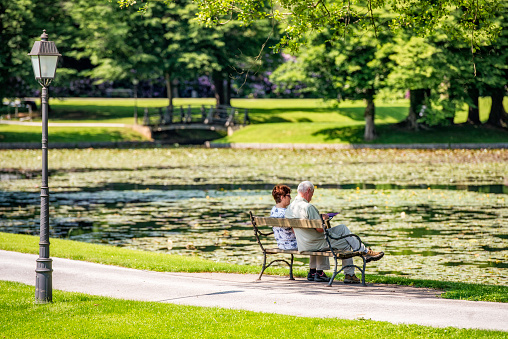 Rear view of a senior couple sitting and relaxing next to a lake in a public park on a sunny day.