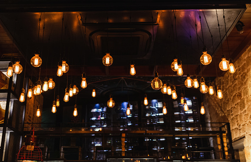 Many electrical lamps bulbs hanging from ceiling at restaurant interior and glowing in darkness. Dark background. Horizontal color photography.