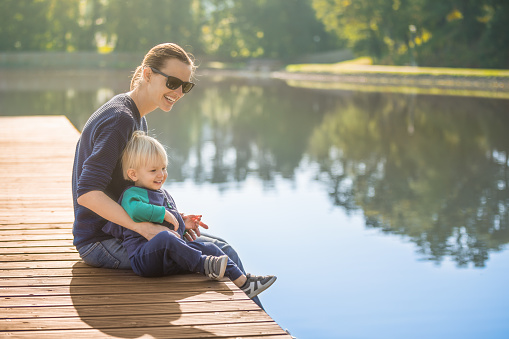 Happy smiling mother and toddler son relaxing on a wooden dock lake side.