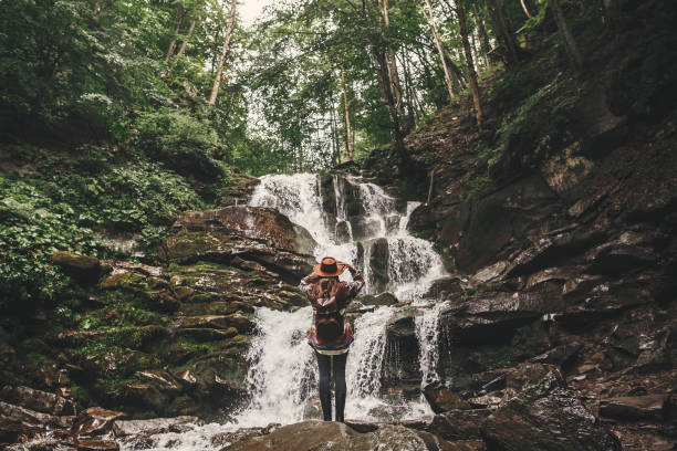 istock Young woman with backpack exploring in summer forest, looking at waterfall. Travel and wanderlust concept. Space for text. Stylish hipster girl in hat traveling in mountains. 1089240488