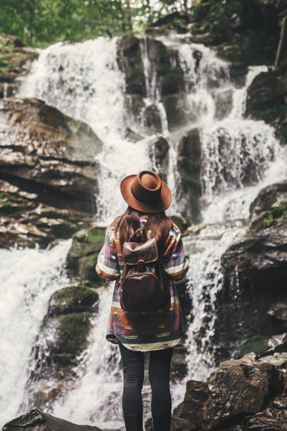 istock Young woman with backpack exploring in summer forest, looking at waterfall. Travel and wanderlust concept. Space for text. Stylish hipster girl in hat traveling in mountains. 1089240890