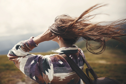 Stylish hipster girl with backpack  and windy hair on top of mountains. Portrait of happy young woman relaxing. Carefree mood. Amazing atmospheric moment