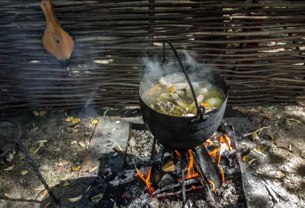 Chicken soup in a pot is cooked on an open fire near a wicker fence. Rural environment, country life. Evening light, the rays of the setting sun, sunset.