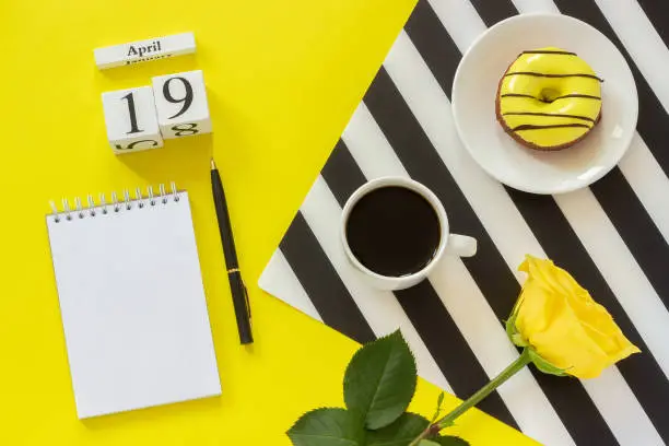 Photo of Wooden cubes calendar April 19th. Cup of coffee, yellow donut and rose on black and white napkin, empty open notepad for text on yellow background. Concept stylish workplace
