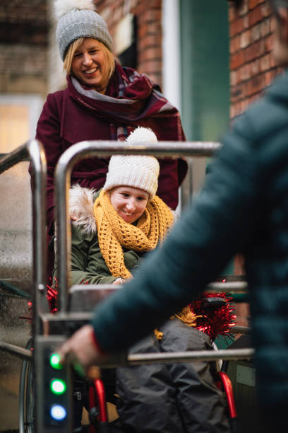 Family Using Wheelchair Lift A front-view shot of a mother and father helping their physically impaired daughter in her wheelchair, they are wearing warm clothing as they exit a building using a wheelchair lift. wheelchair lift stock pictures, royalty-free photos & images