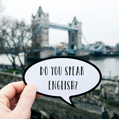 closeup of a young caucasian man holding a signboard in the shape of a speech bubble, with the question do you speak English? written in it, in front of the Tower Bridge in London, United Kingdom