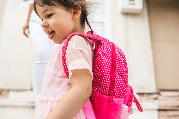 cropped closeup image of beautiful kid showing tongue and smiling as she leave for kindergarten against her mother. cute happy little girl wears backpack going to preschool. education, people concept - primary care imagens e fotografias de stock