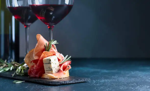 Sandwich with prosciutto, blue cheese and rosemary on a dark background. Glasses and bottle of red wine with snack. Copy space for your text.