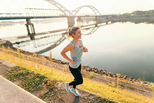 Happy sportive blonde woman in sportswear running by the river in the morning.