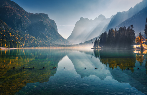 Majestic mountains reflected in water in beautiful Dobbiaco lake at sunny morning in autumn.  Fall in Dolomites, Italy. Landscape with hills, trees, blue water and bright sunlight at sunrise. Nature