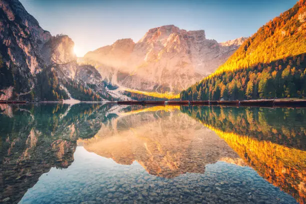 Beautiful Braies lake at sunrise in autumn in Dolomites, Italy. Landscape with mountains, gold sunlight, water with reflection, trees with orange leaves in fall morning. Travel in italian alps. Nature