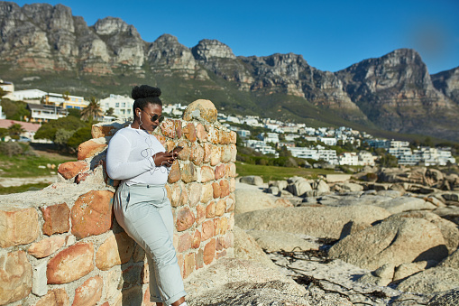 Young woman using mobile phone while leaning on stone wall. Female is listening music with town and mountains in background.