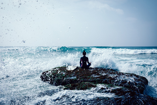 Woman surfer meditation at the seaside coral reef edge facing the coming strong sea waves