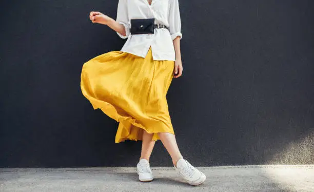 Photo of Horizontal cropped image of stylish slim woman in beautiful yellow skirt. Caucasian female fashion model standing over gray wall background outdoor with copy space.